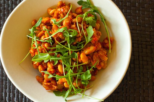 white beans cooked in red sauce in a white shallow bowl, garnished with arugula